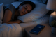 Person sleeping in bed with a smartphone on a bedside table emitting symbolic wireless signal waves