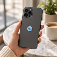 EMF Mini Shield – Glitz Cool Breeze Blue on the back of a smartphone, held in hand at a café — glitter light-blue EMF phone protection sticker
